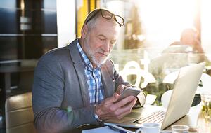 Senior businessman sits at café table working on laptop and looking at smartphone